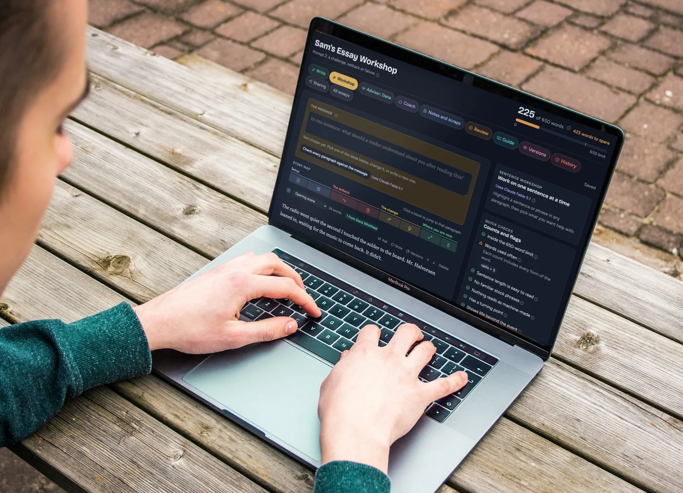 Over the shoulder of a student typing on a laptop at an outdoor table. The screen shows the AdmitUs workshop in dark mode with a made-up example essay.